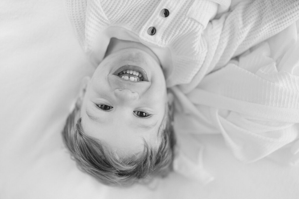 Black and white image of a toddler boy looking at the camera as he laughs rolling around on a bed during a session with Carmel family photographer, Brittney Lear Photography