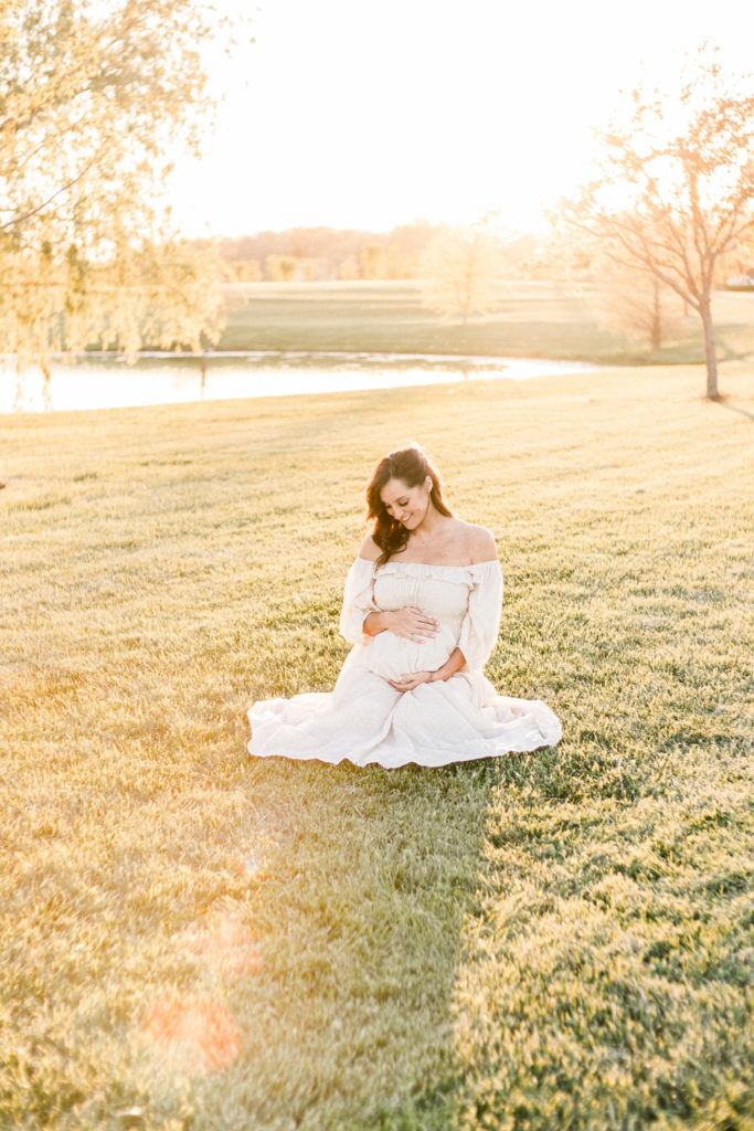 7 months pregnant woman sitting by pond and smiles in light of the sunset at maternity session at coxhall gardens in carmel indiana