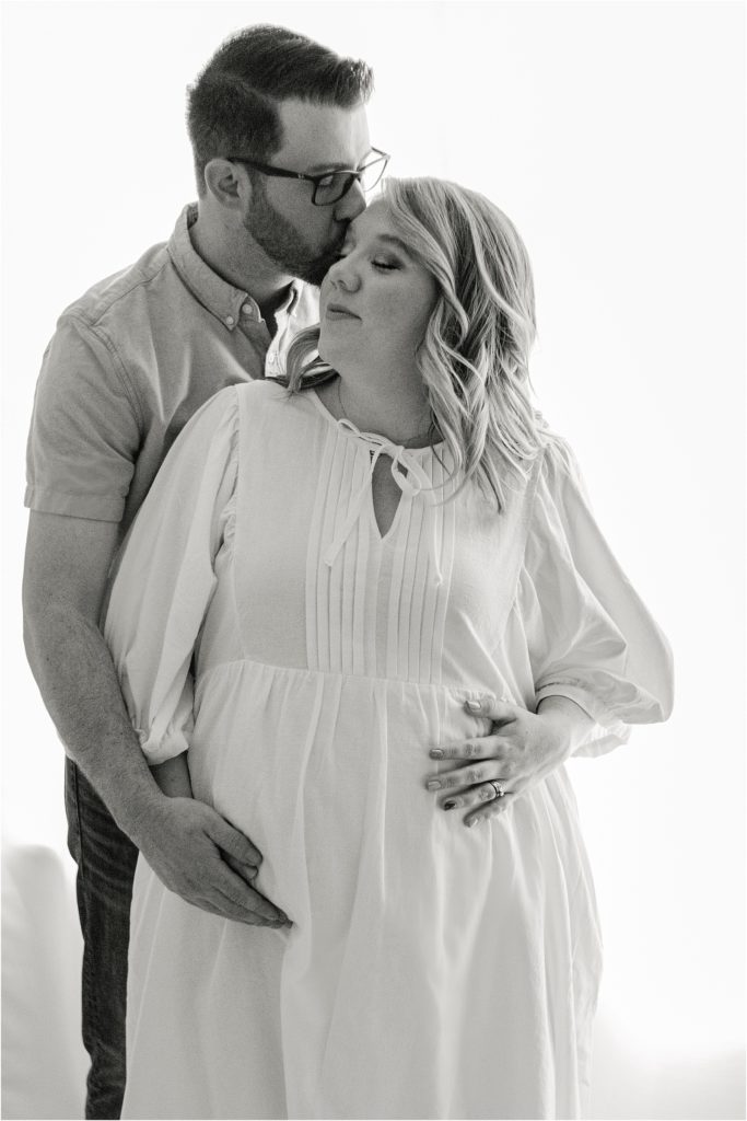 couple holding baby bump while husband kisses forehead