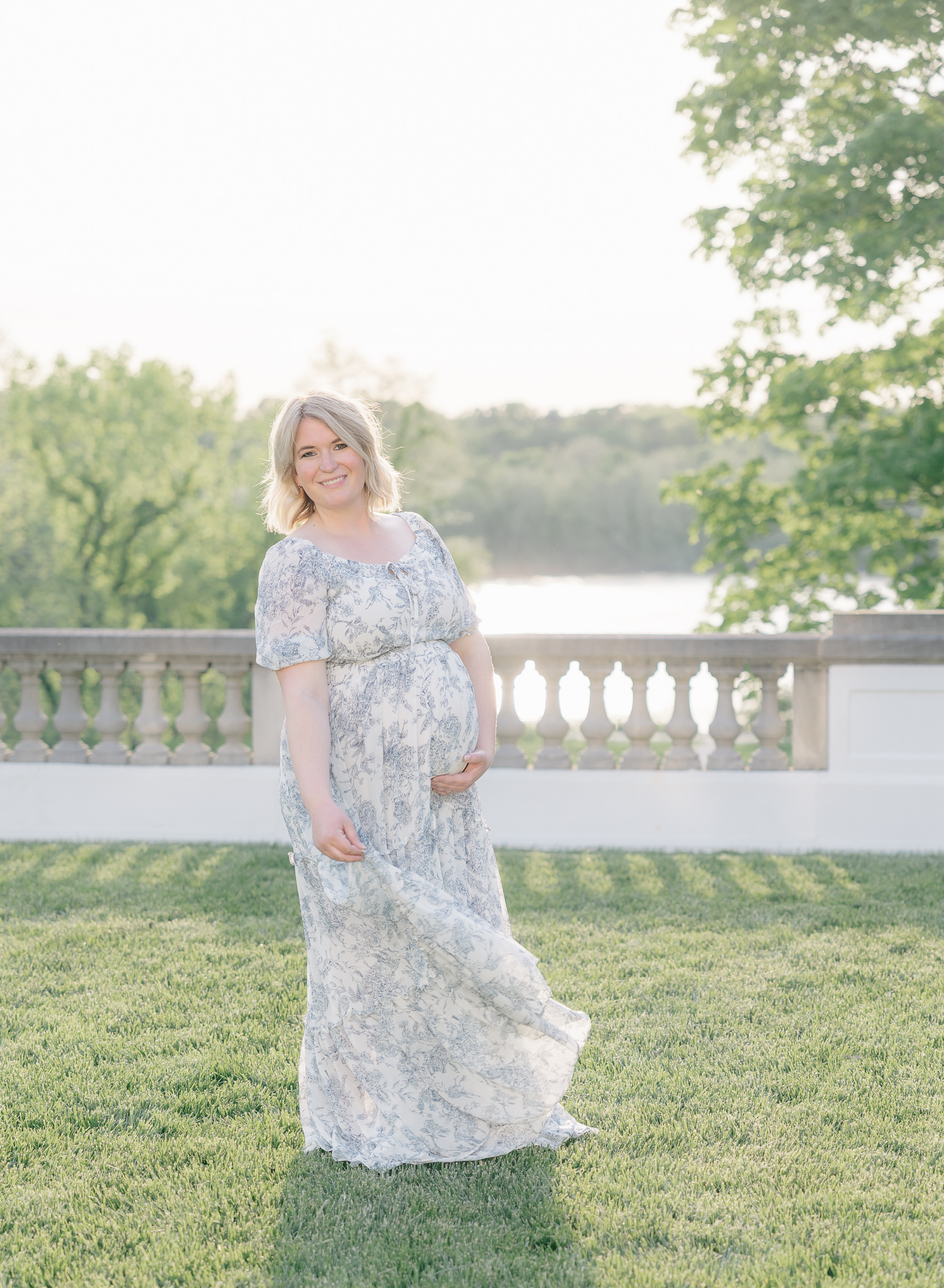 mother in white and blue floral dress holding baby bump while standing on the terrace at the Lily House at sunset, Session styling tips for expecting mothers, Zionsville maternity photographer
