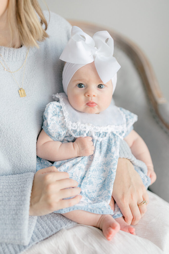 mother in blue sweater holding baby girl in blue heirloom dress and large white bow as baby looks into camera, Carmel Family photography