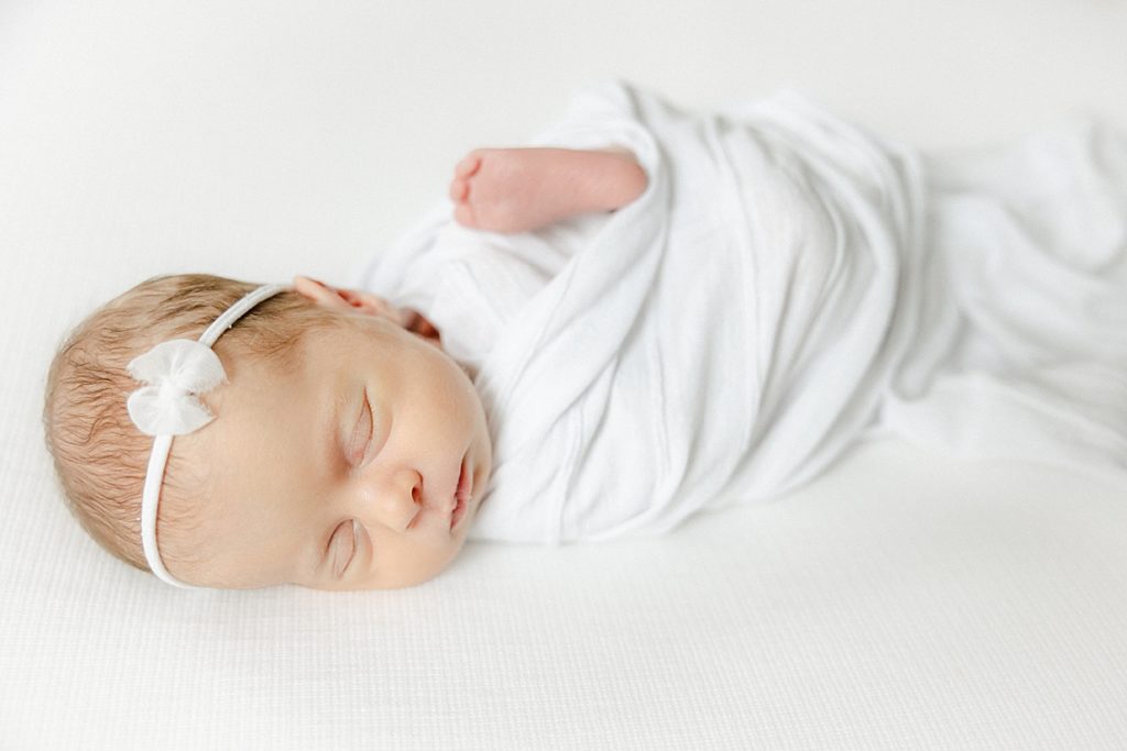 Newborn baby girl sleeping on a white blanket swaddled in white with her foot out Carmel newborn photographer