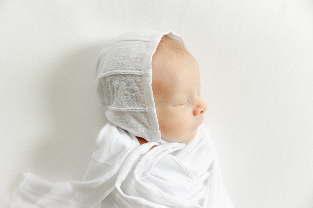 Side profile of a newborn baby boy sleeping on a white blanket while wearing a white bonnet 