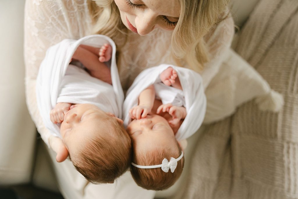 Mother holding her twin babies swaddled in white while looking down towards them as they find their hands Carmel Newborn photographer