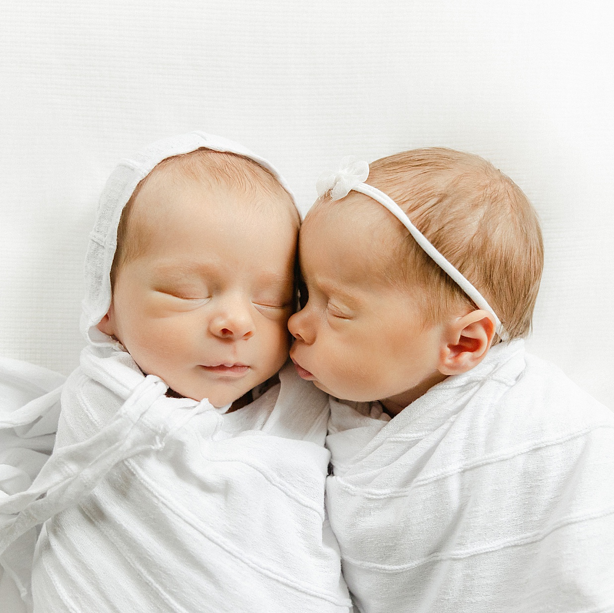 Baby girl leaning in to kiss her twin brother's cheek while swaddled in white on a white blanket during their session with a Carmel newborn photographer