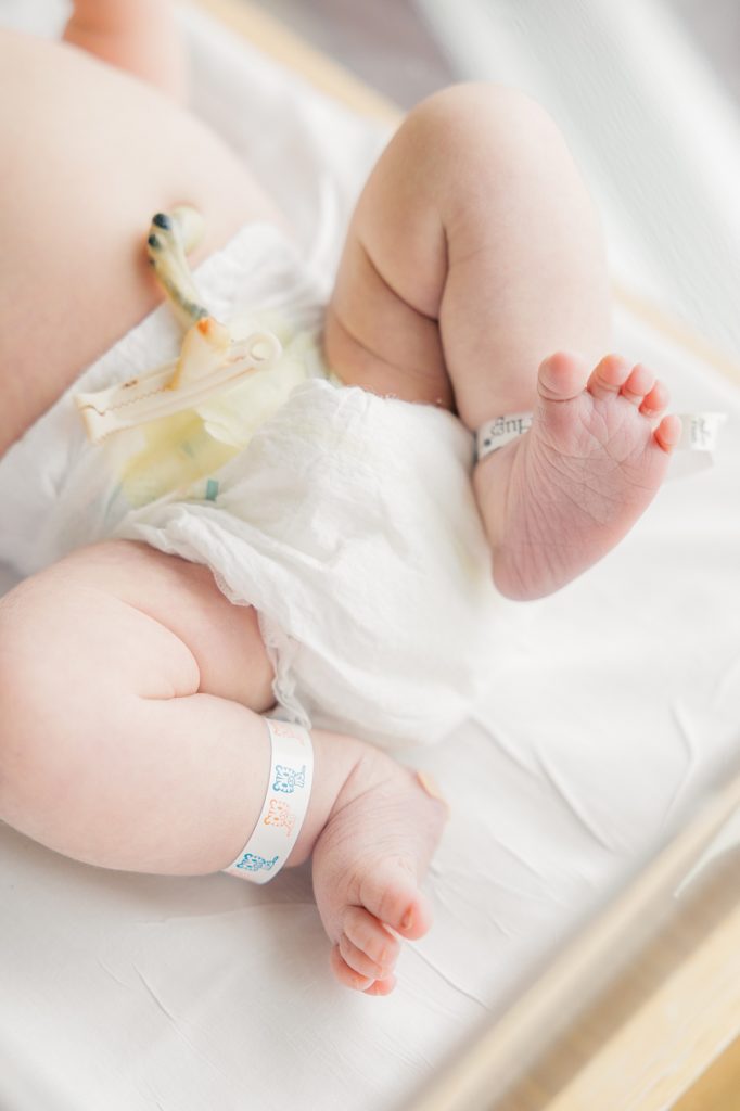 Newborn baby wearing diaper and hospital bracelets on ankles holds one leg up in the air on the hospital bed