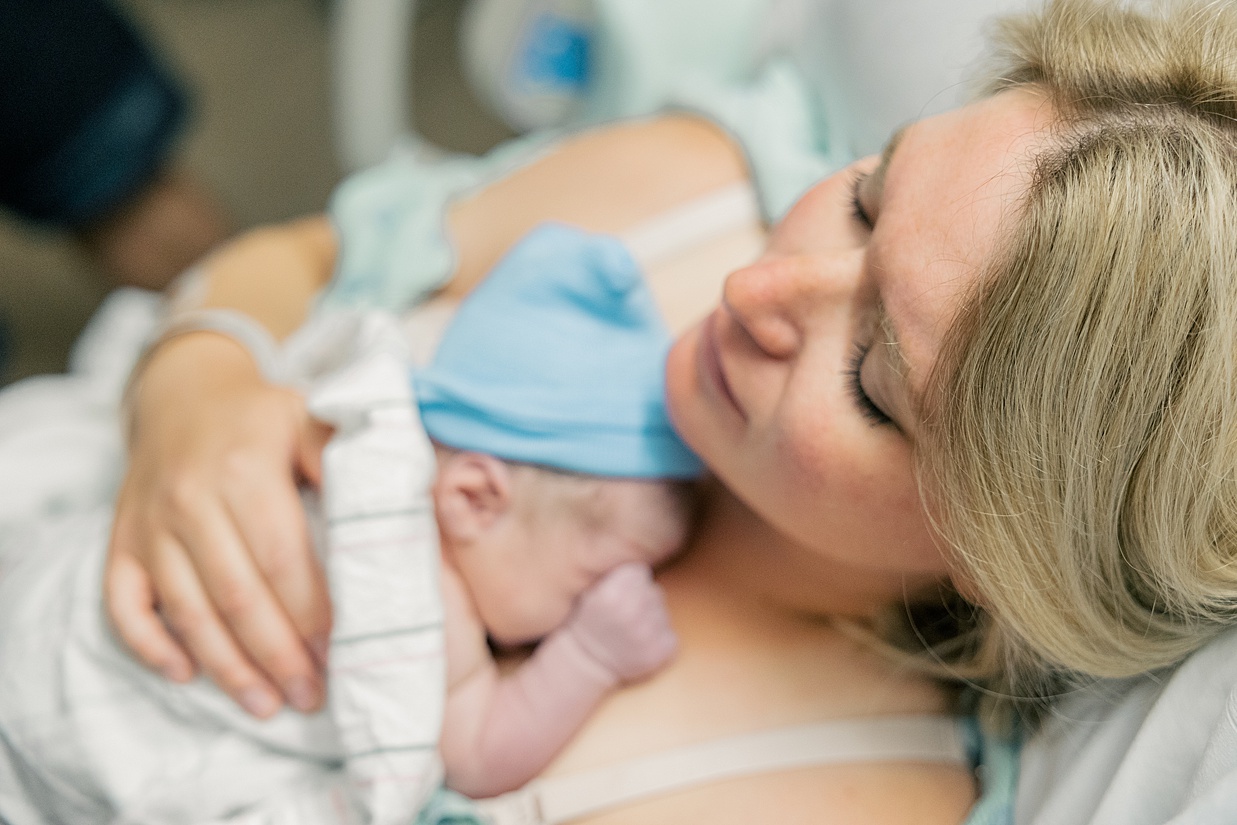 Mother laying in hospital bed with newborn baby on her chest wearing blue hat and white blanket after a smooth delivery thanks to birthing classes in Indianapolis