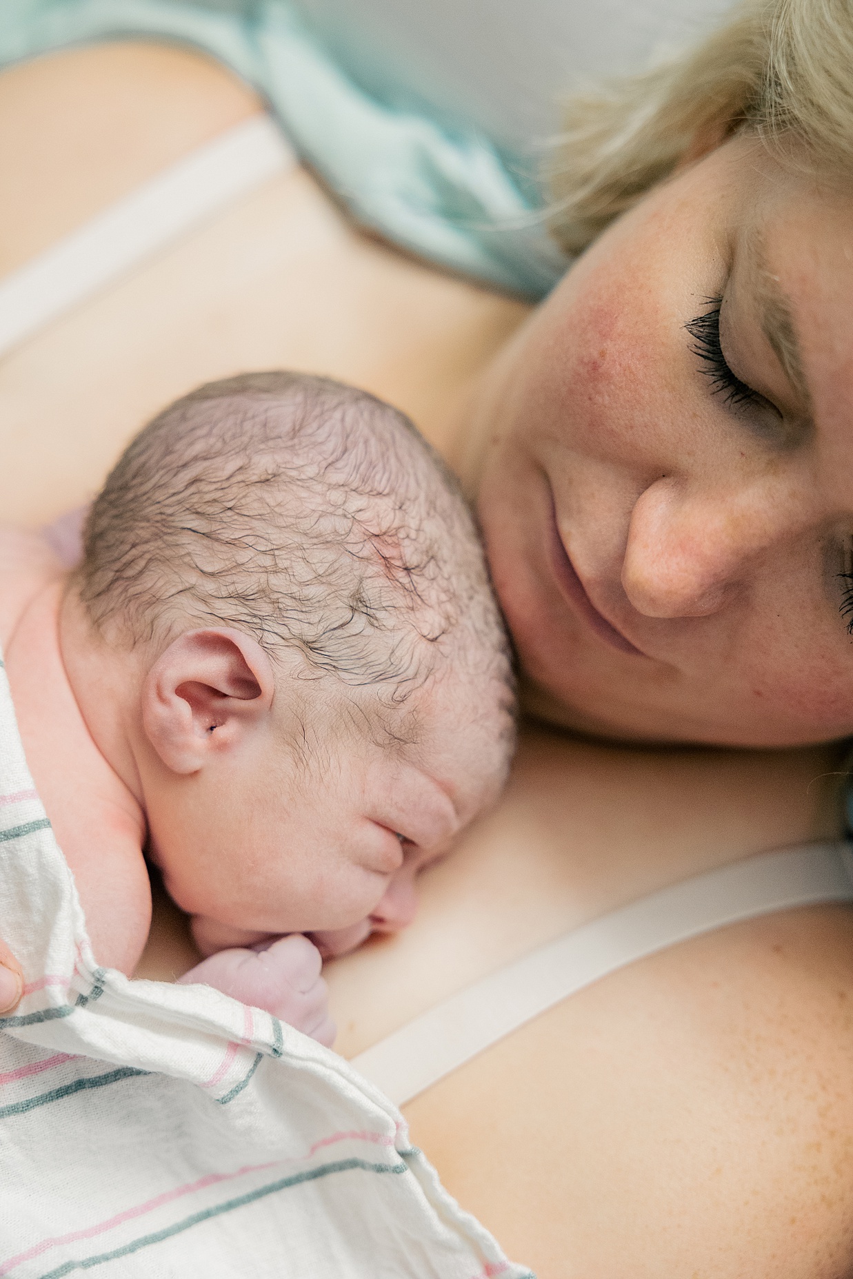New mother holds her newborn baby on her chest while laying in bed with white blanket