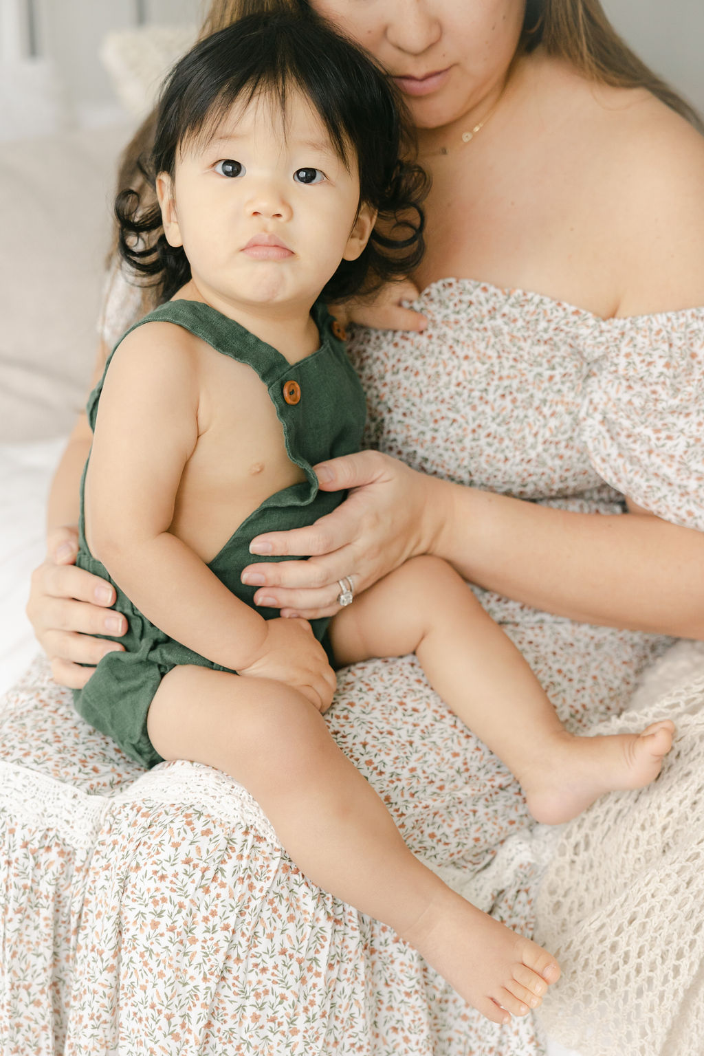 toddler in green overalls sits on mother's lap on a bed Indianapolis baby boutique