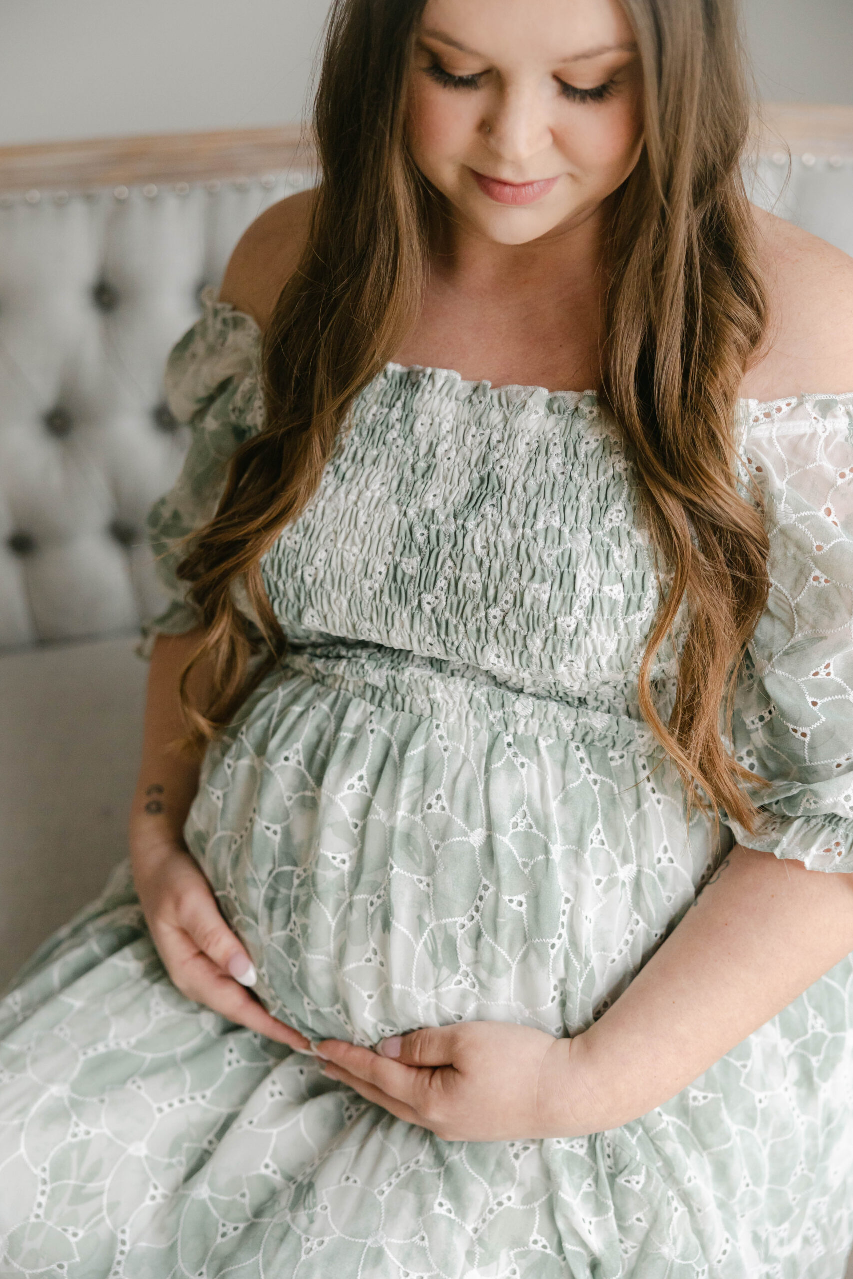 Mother holding baby bump while sitting on grey loveseat in sage floral dress, second trimester, session styling tips for expecting mothers, Zionsville maternity photographer
