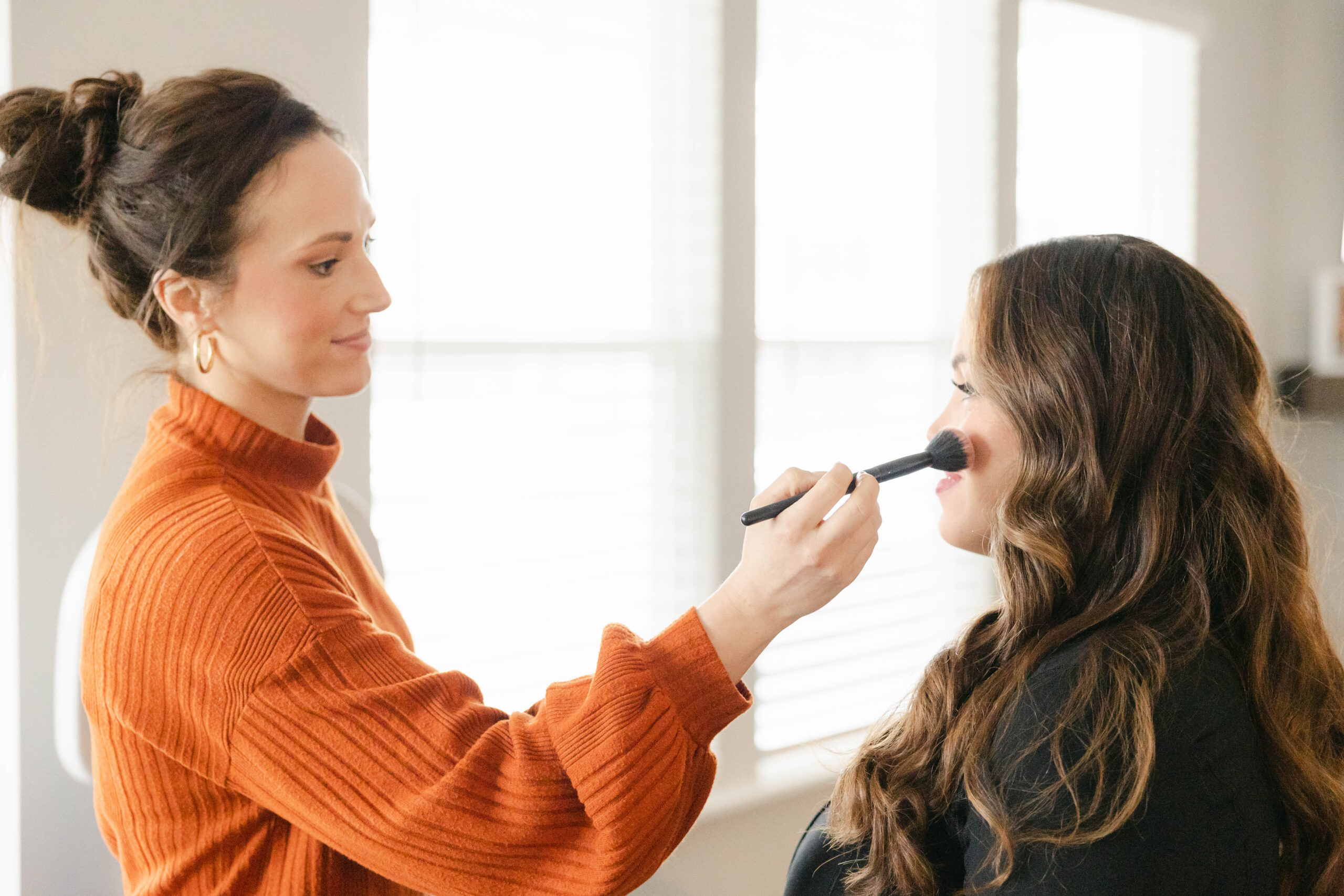 Expecting mother sitting in front of large windows while a makeup artist applies blush with a brush to her face, Zionsville maternity photographer