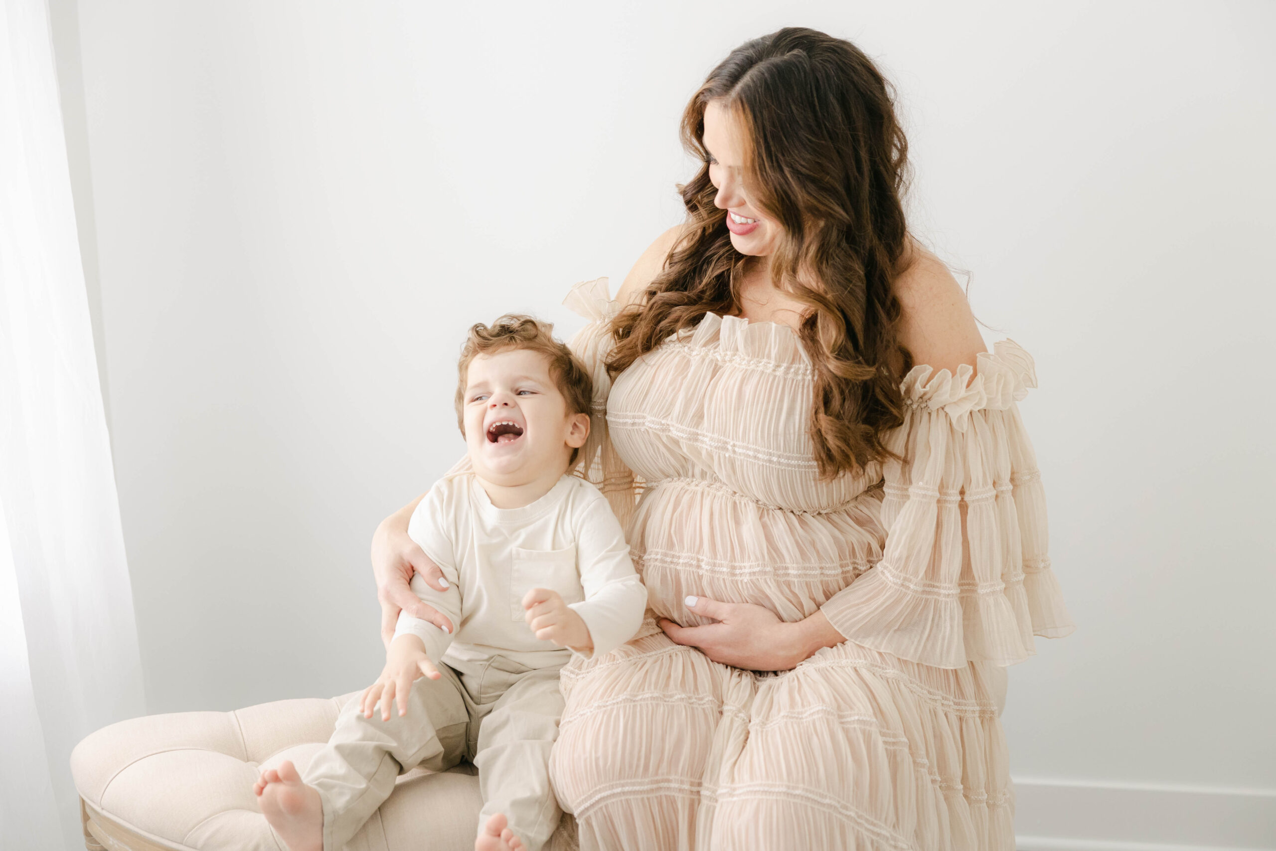 mother and toddler boy in cream outfits sit on a bench snuggled close as the little boy throws his head back in laughter