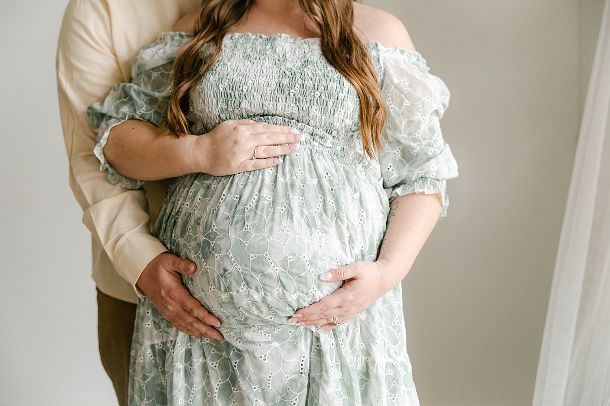 Expecting mother in a sage floral dress places her hands on her baby bump while her husband in a cream shirt places his hand on her bump as well, Maternity photography in Zionsville