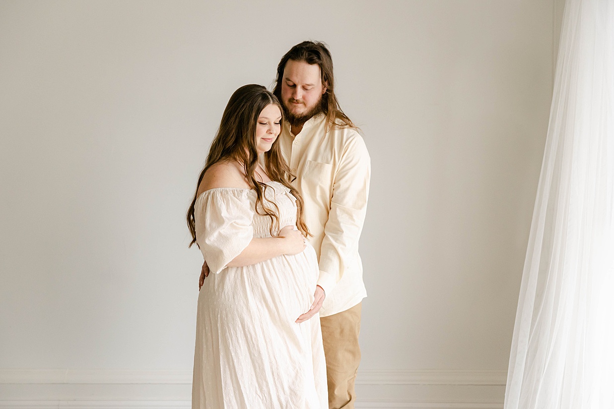 Expecting mother in a cream dress holds her baby bump while her husband feels for baby's kicks, Maternity photography in Zionsville