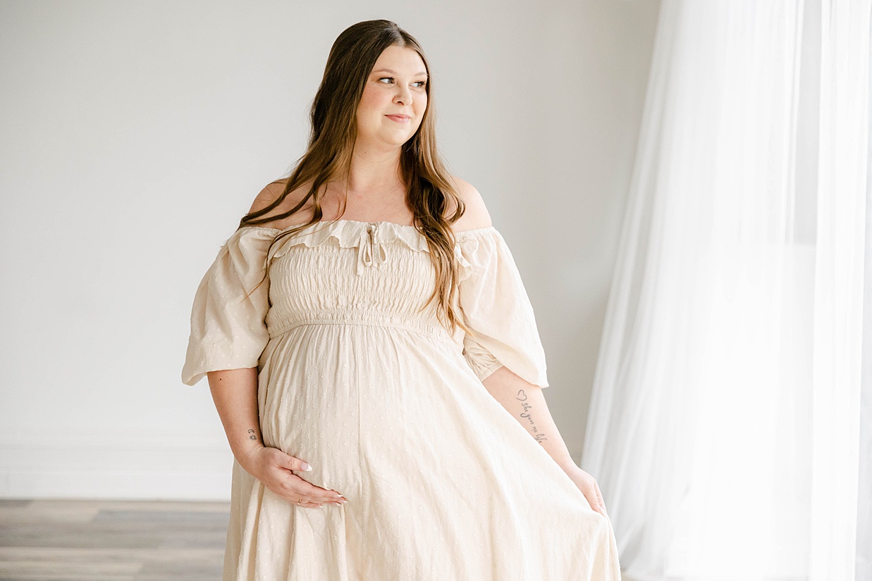 Pregnant mother in a cream dress walks while holding her baby bump while looking out the window, Maternity photography in Zionsville