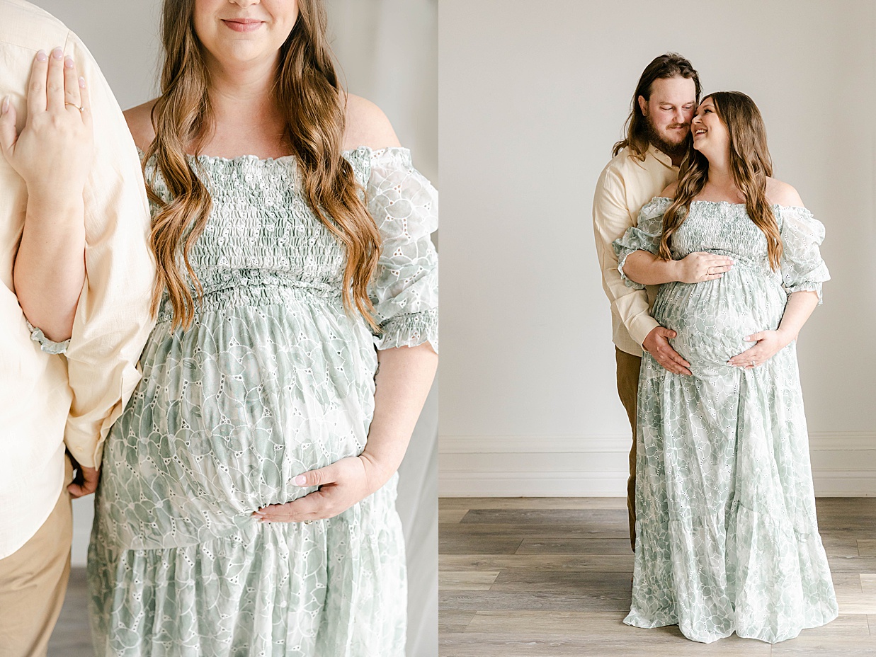 Expecting mother in a sage floral dress places her hands on her baby bump while looking at her husband in a cream shirt standing behind her with his hand on her bump as well, Maternity photography in Zionsville