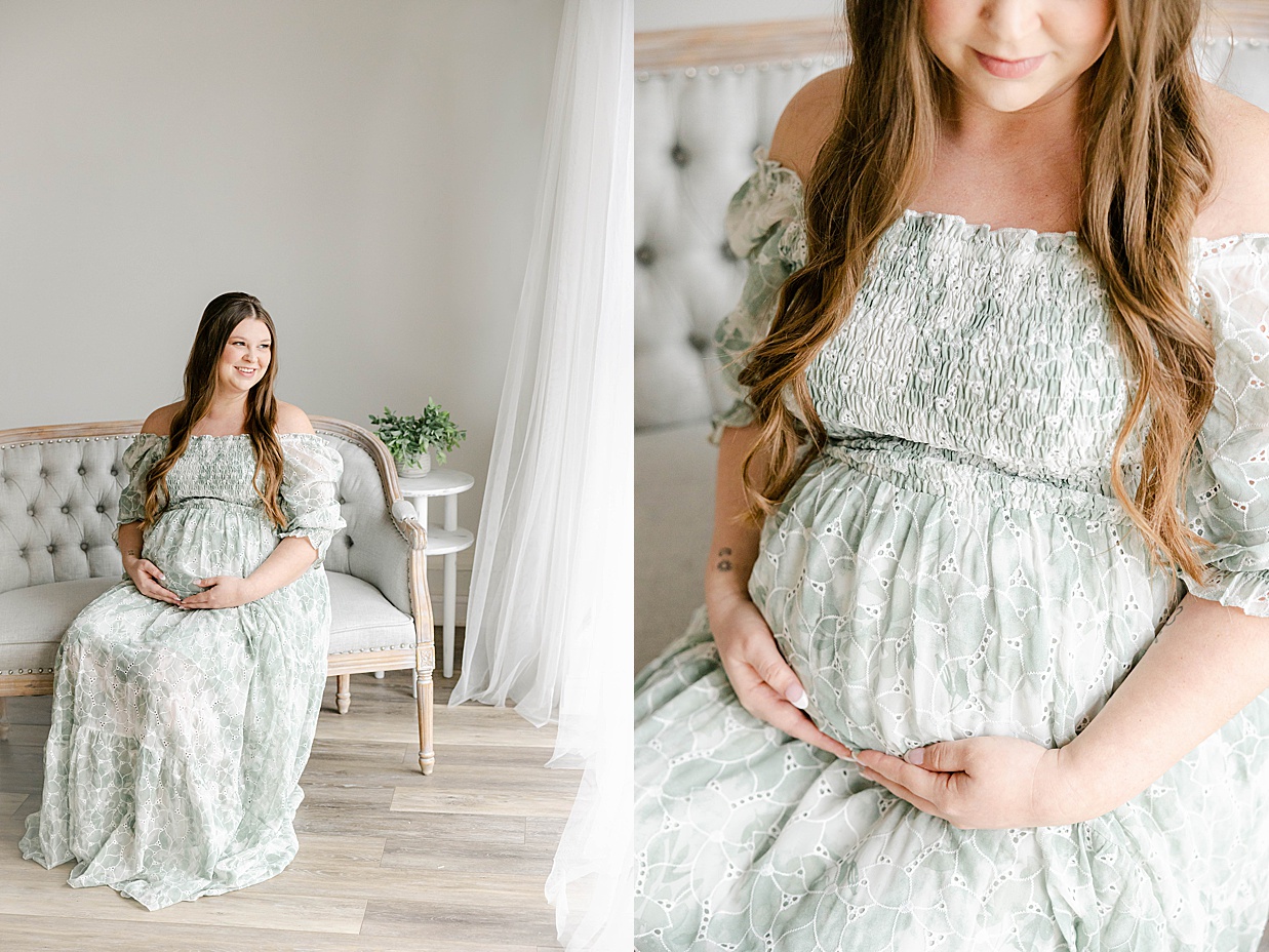 Pregnant mother in a sage green dress holds her baby bump while sitting on a grey loveseat and looking out the window, maternity photography in Zionsville