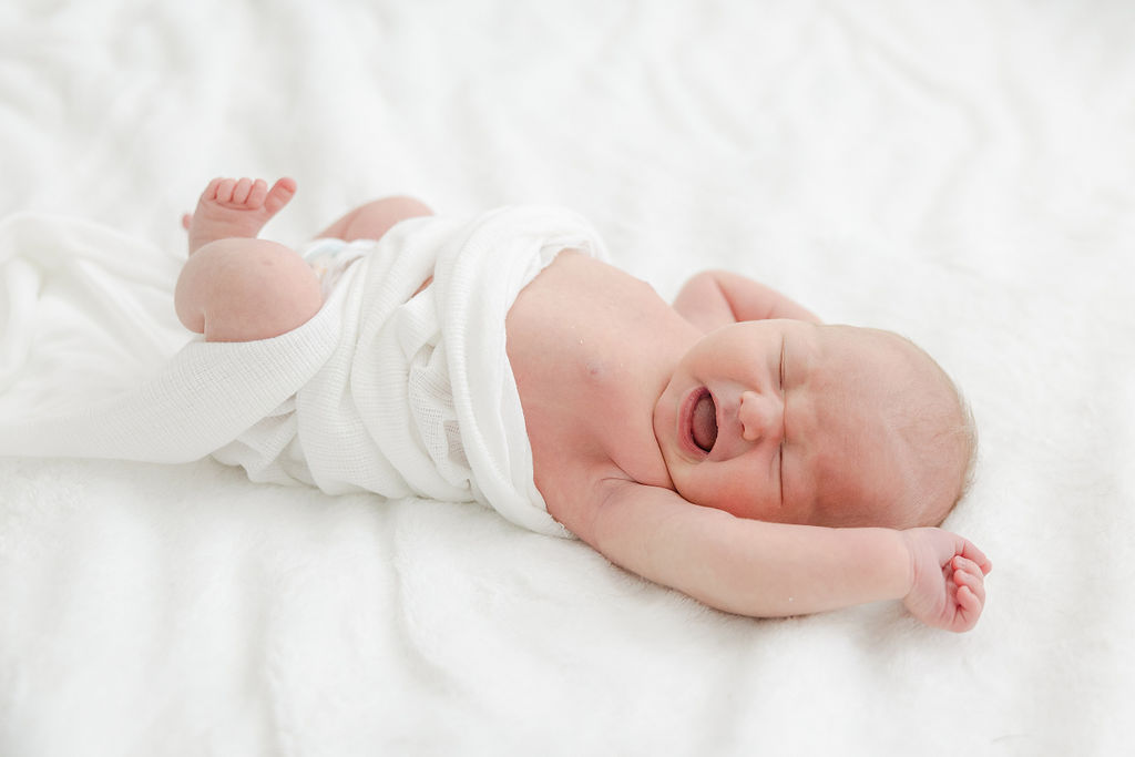 A newborn baby lays on a white bed with a white blanket wrapped around its waist with arms stretched up and mouth open indianapolis pediatricians
