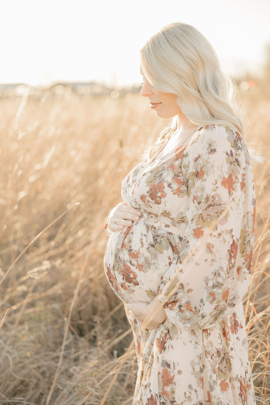 A pregnant woman looks down on and holds her bump while standing in a field of golden grass at sunset in a floral pattern maternity dress indianapolis prenatal massage