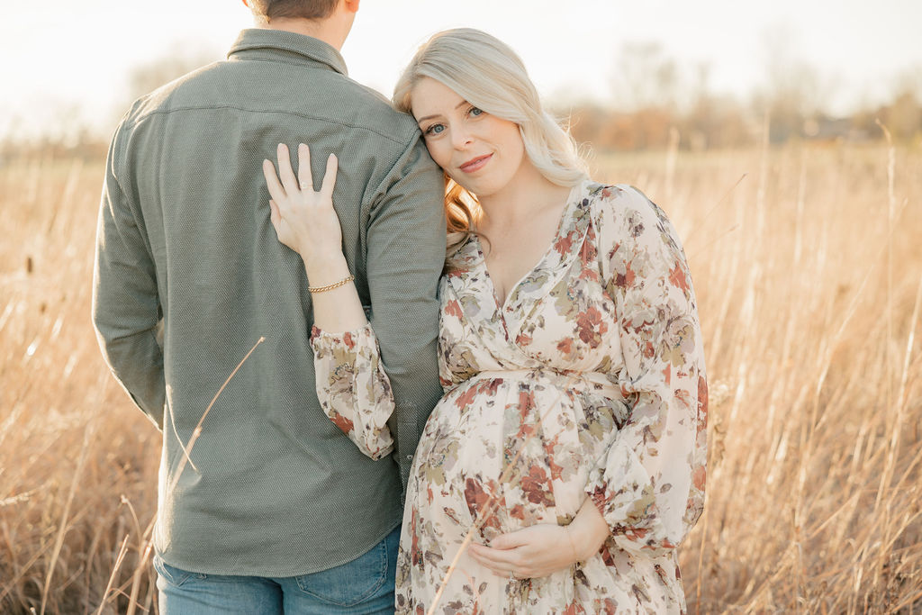 A mother-to-be and her husband stand in a field of tall golden grass while holding her bump in a floral maternity dress indianapolis prenatal massage