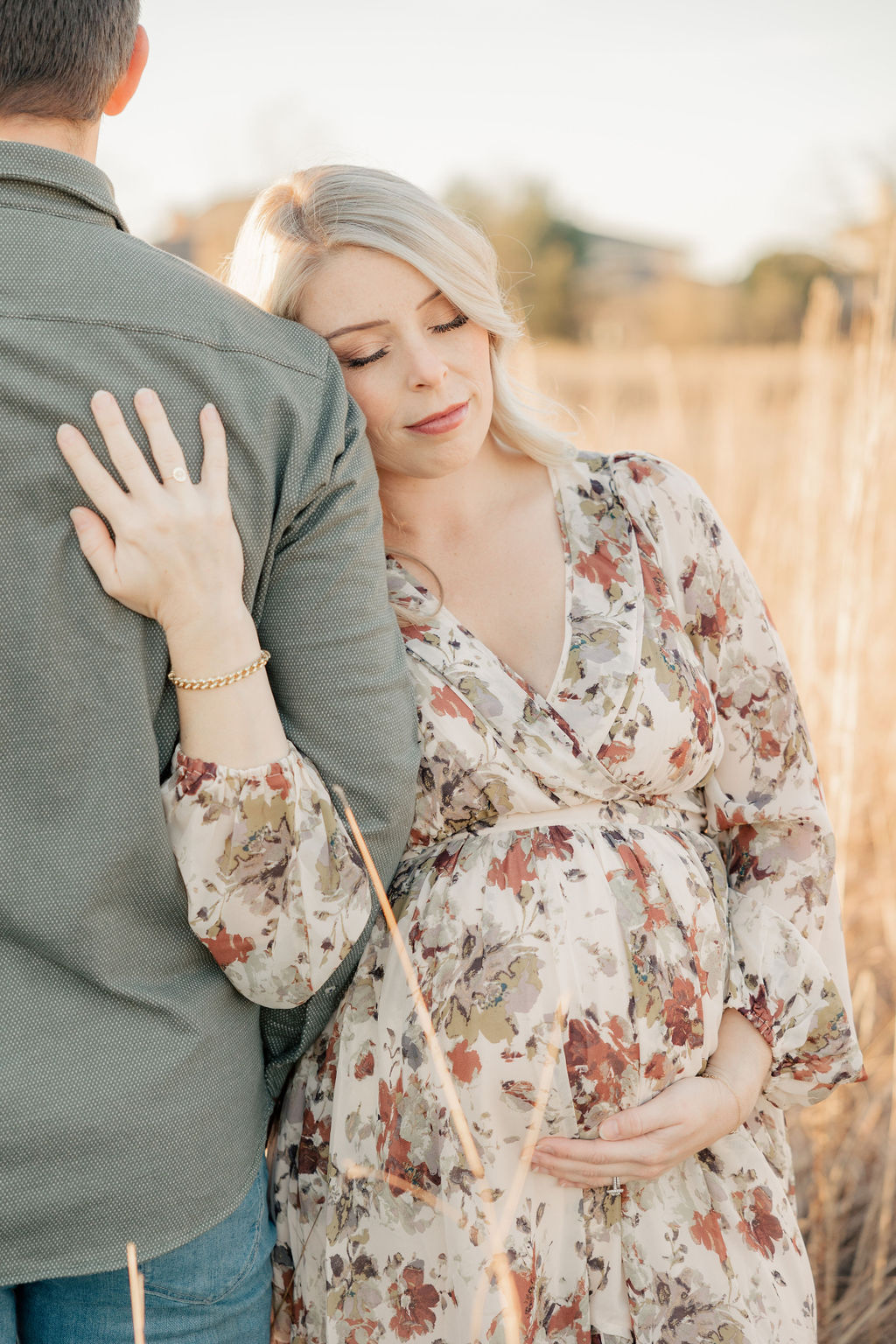 A mother-to-be in a floral pattern maternity gown leans onto her husband's shoulder in a golden field indianapolis prenatal massage