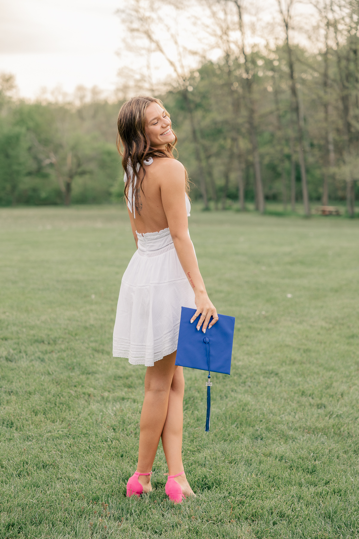 girl in a short white dress and pink heels holds blue graduation cap at the 100 acre woods at Newfields, Indianapolis Senior Pictures