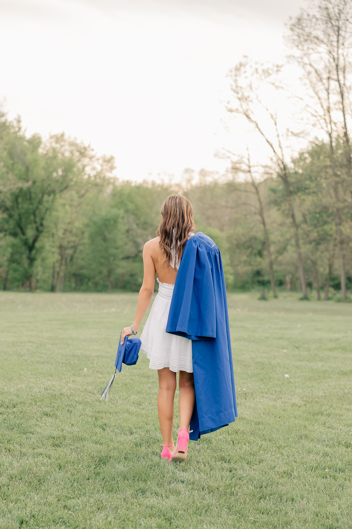 girl in a white dress holds her blue graduation cap and gown while walking away at Newfields, Indianapolis Senior Pictures