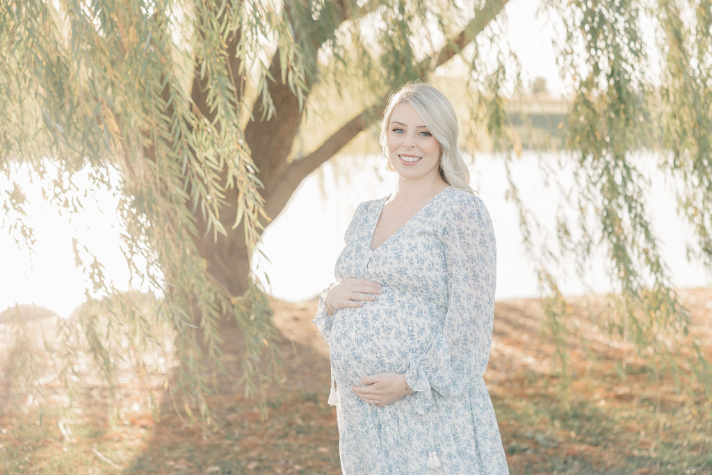 A mother to be stands under a willow tree by water in a maternity dress indianapolis prenatal yoga