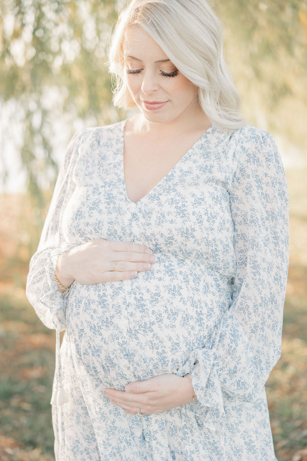A pregnant woman in a white and blue maternity dress stands under a willow tree gazing down at her bump indianapolis prenatal yoga