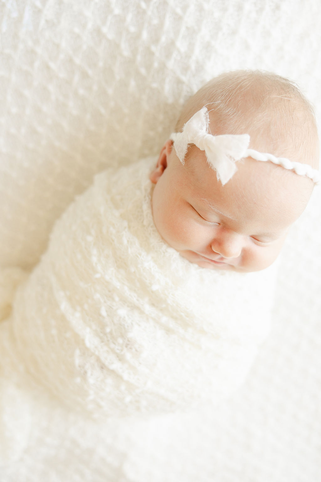 A newborn baby sleeps in a white swaddle with a white bow on her head indy night nanny