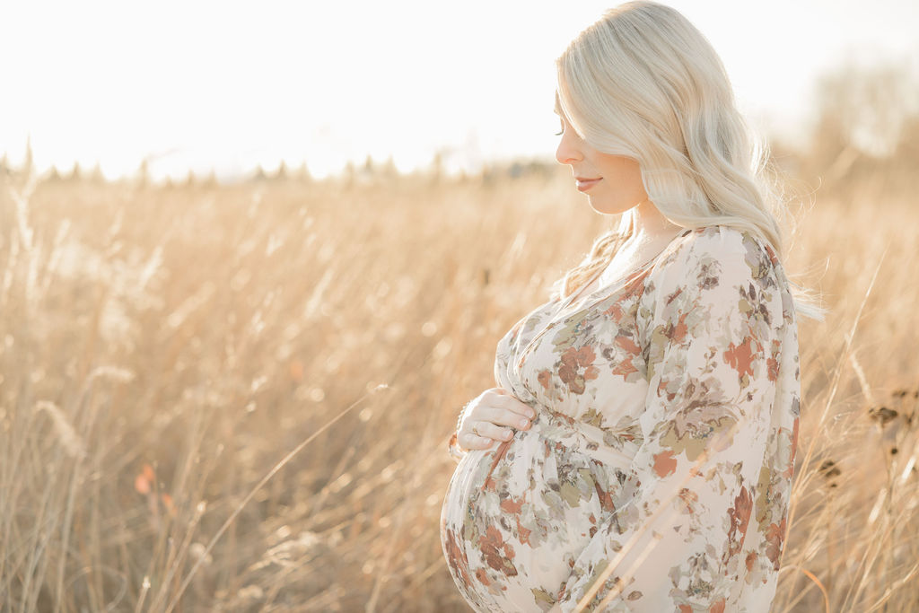 A mother to be in a floral dress stands in a field of flowing golden grass at sunset