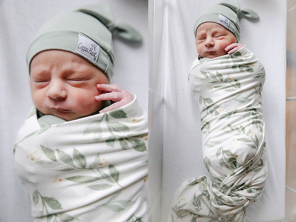 A newborn baby wears a floral swaddle and green sleep cap in a clear hospital crib postpartum doula indianapolis