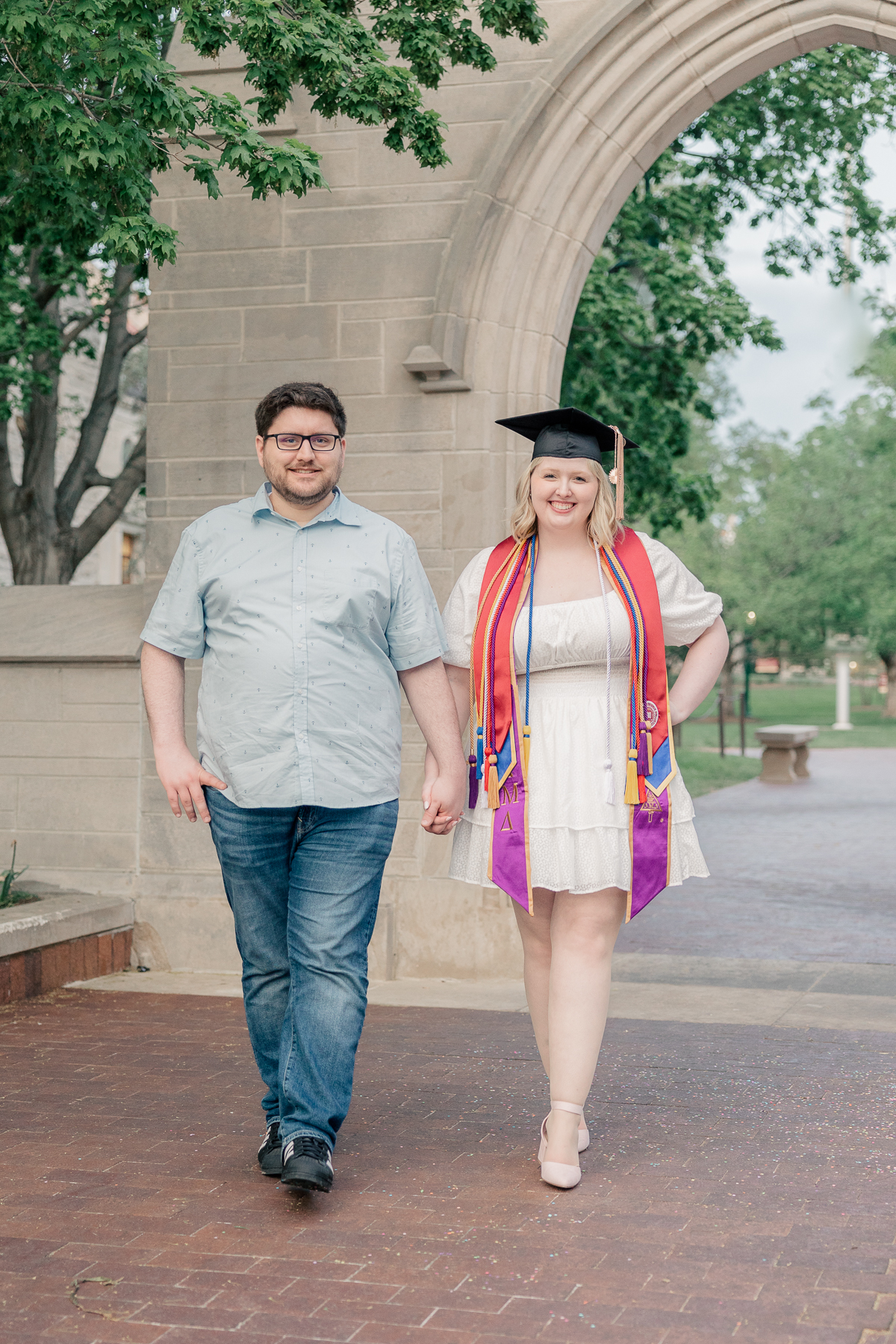 man and woman in white dress and graduation cap walking through stone arch, senior session at IU Bloomington