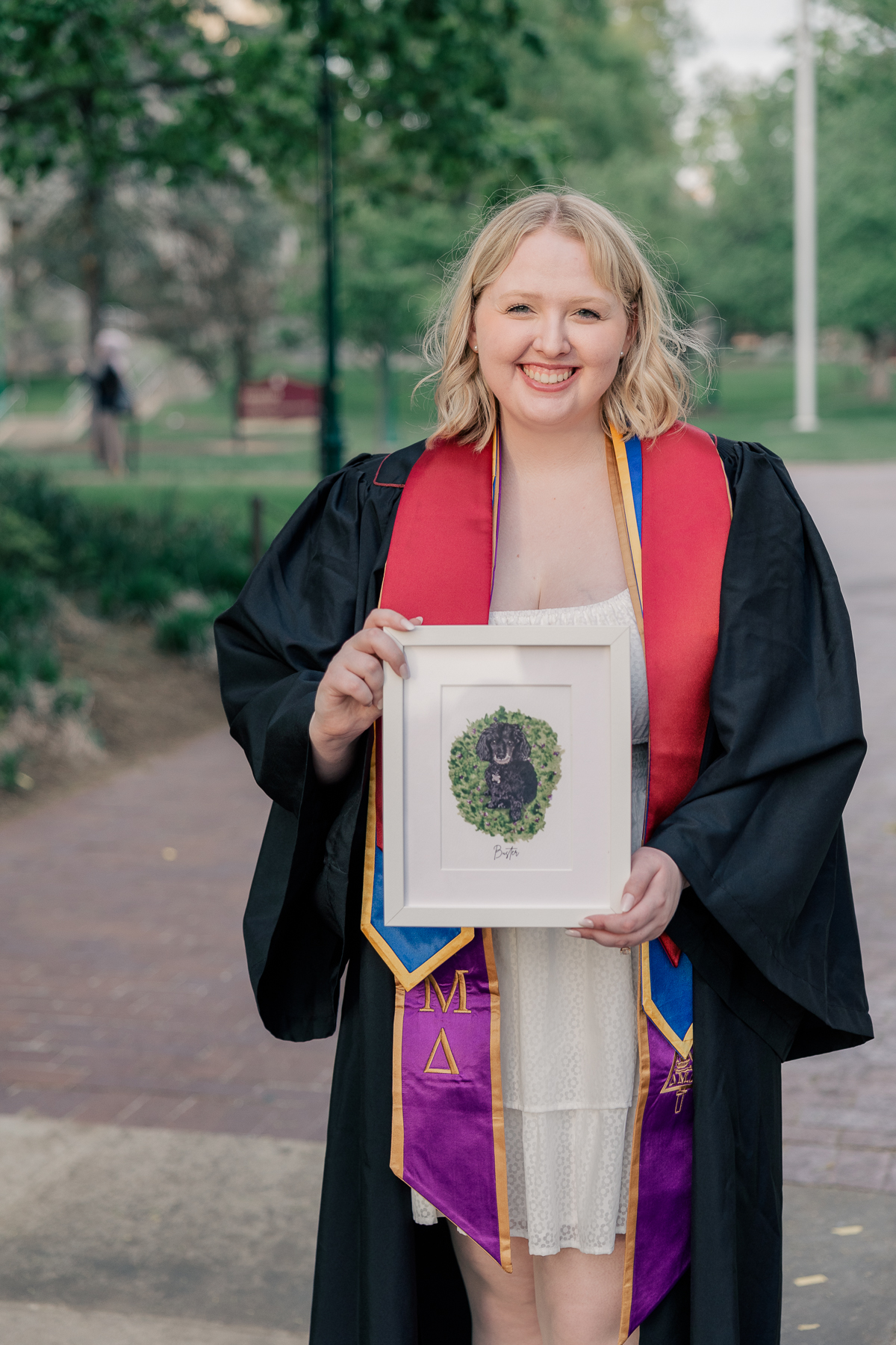 woman in white dress and graduation robes holding a framed image of her dog, senior session at IU Bloomington