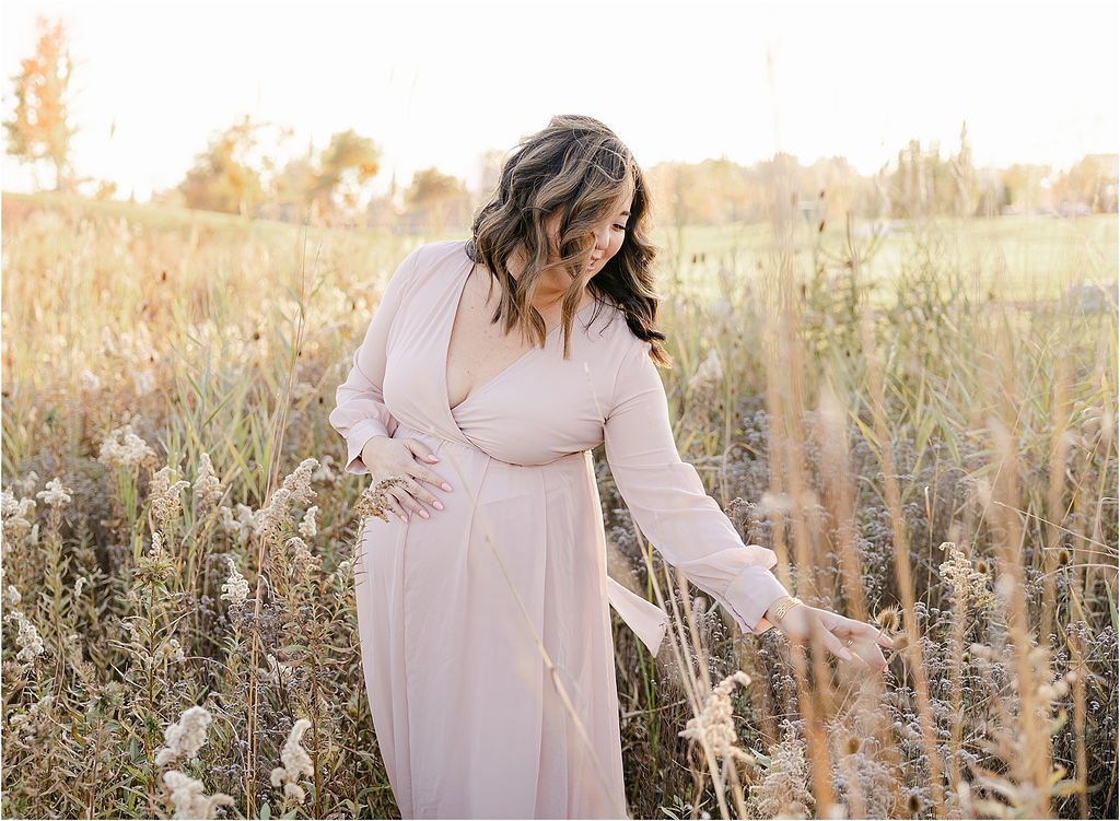 A mom to be in a pink maternity gown walks through and plays with tall grasses in a field
