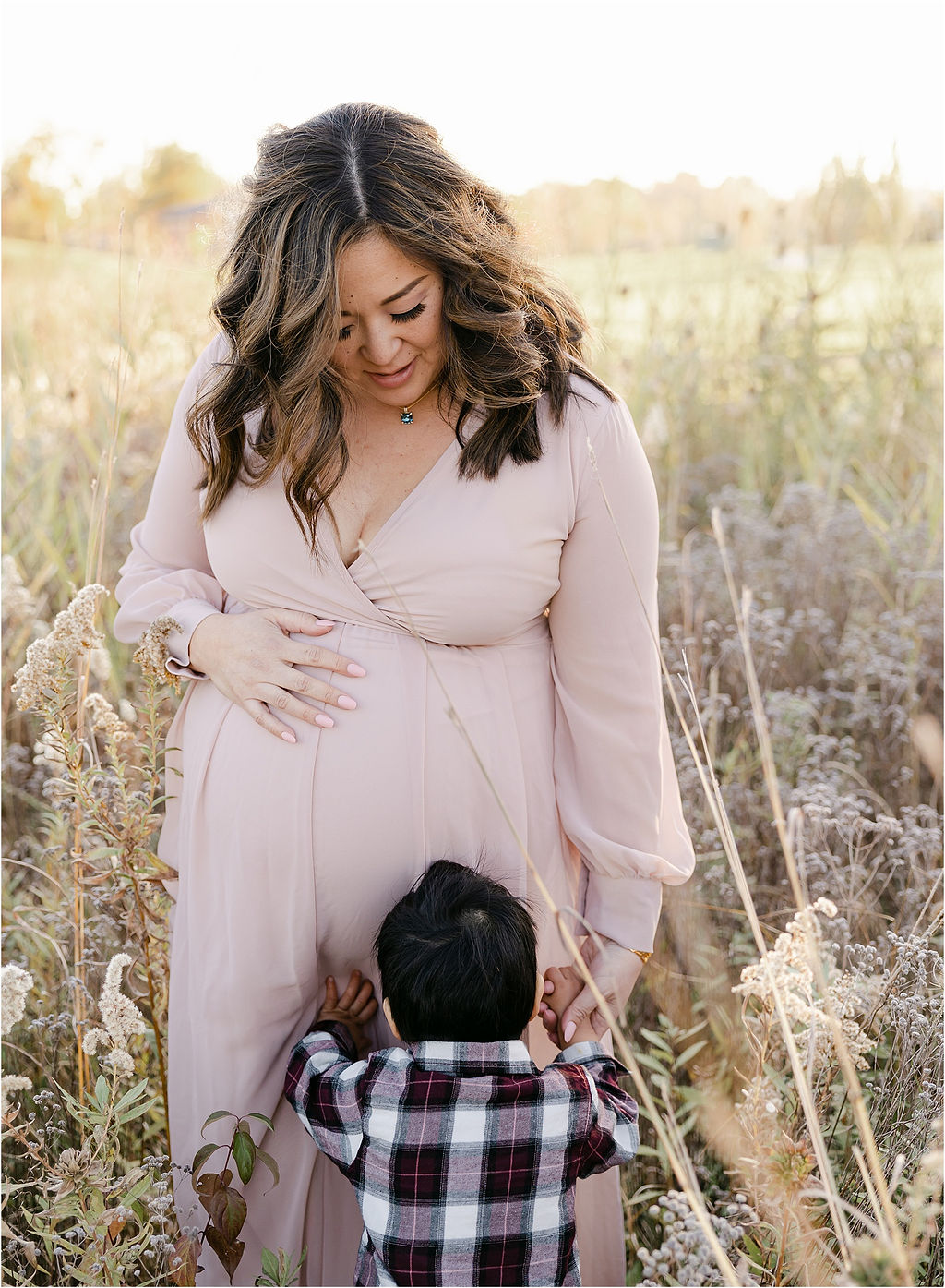 A pregnant mother in a pink maternity gown plays in a field of tall grasses with her toddler son Divine Birth Midwifery