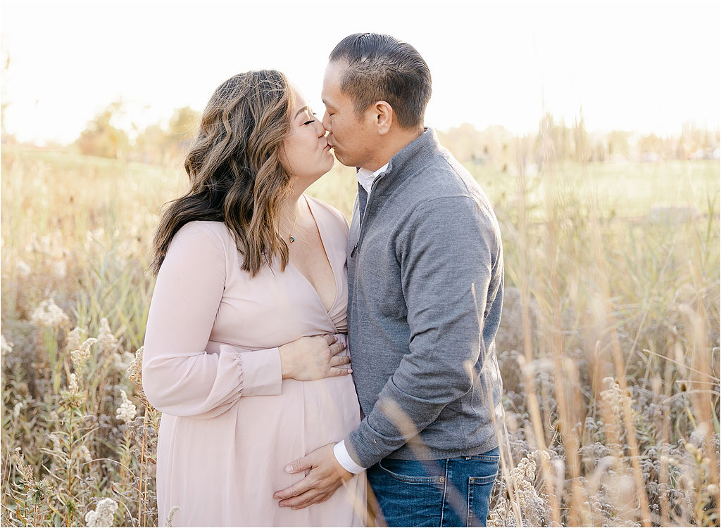 Parents to be kiss while standing in a field of tall grasses with hands on the bump