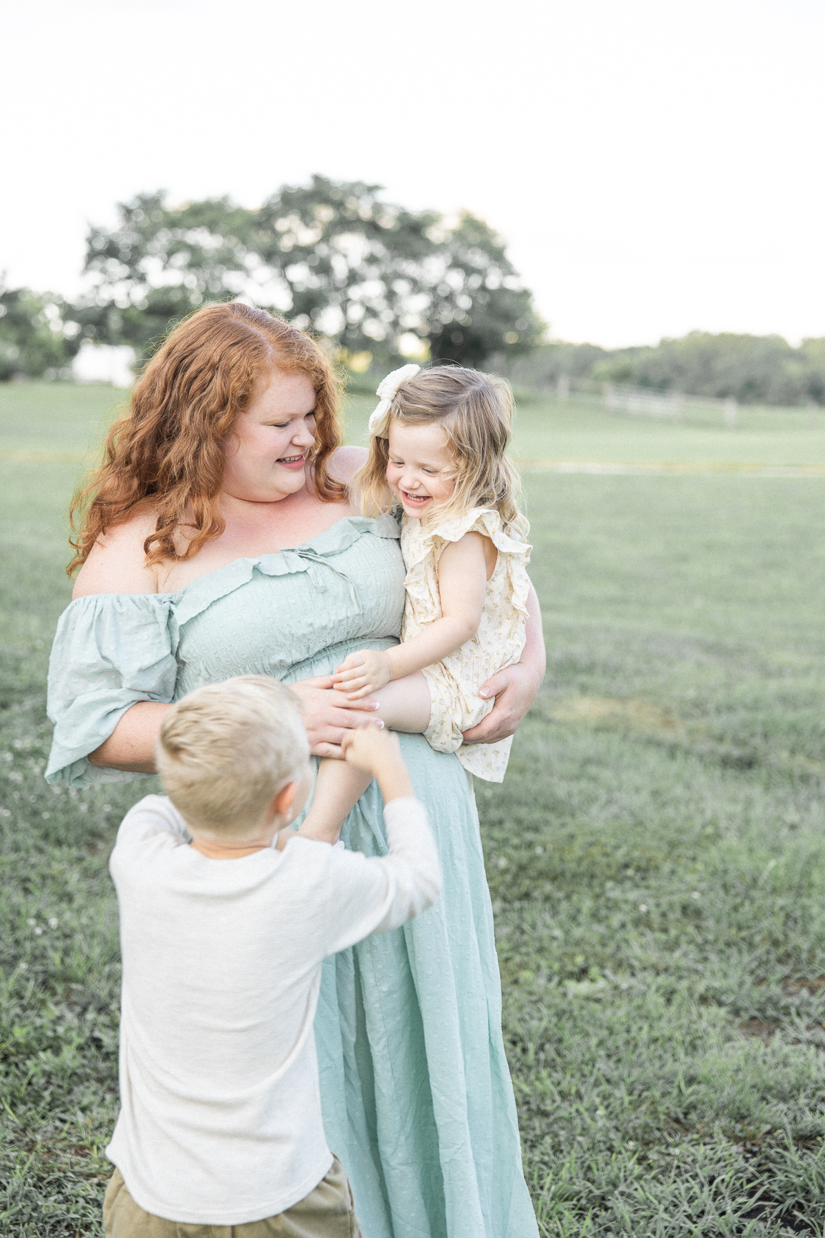 A holds her toddler daughter as her son tickles her leg while standing in a field of grass indianapolis baby shower venues