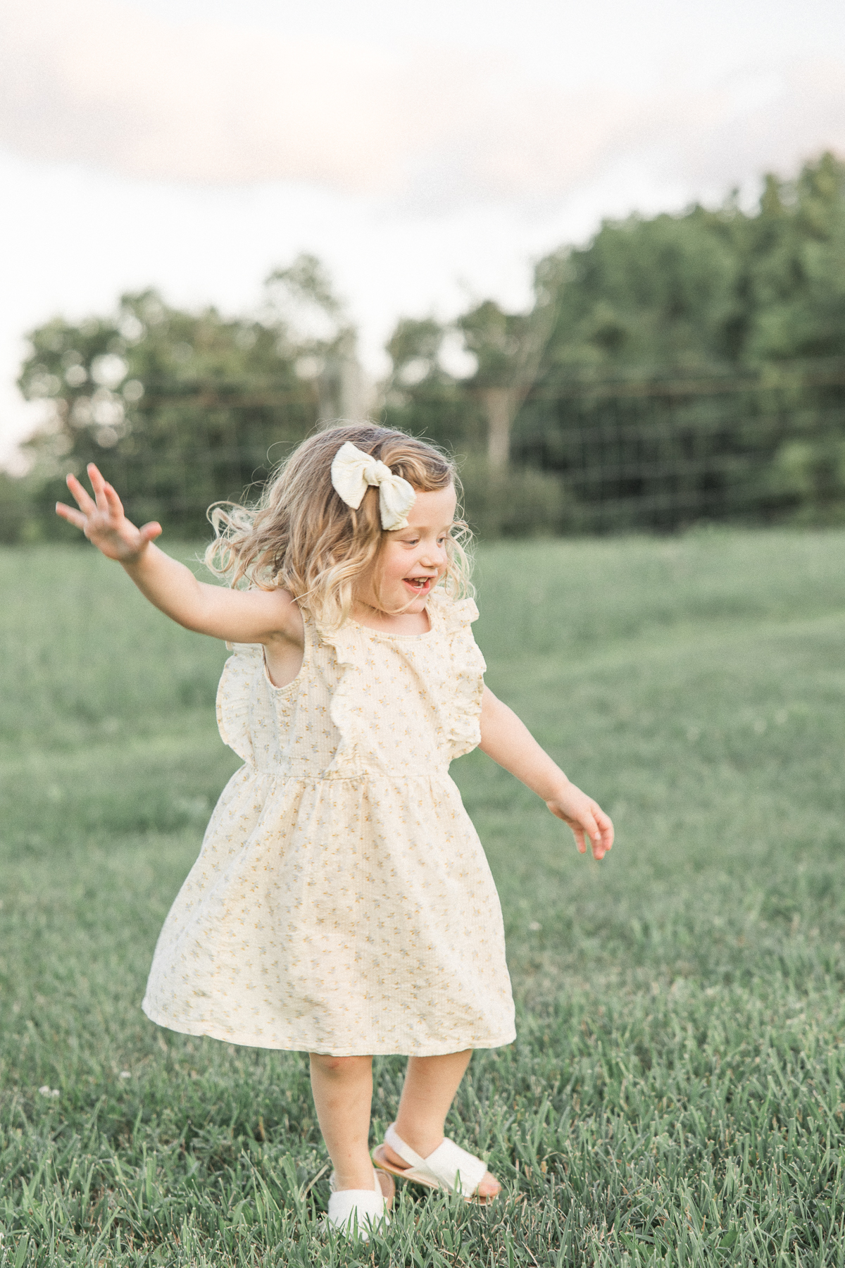young toddler girl with blonde curly hair in cream dress dances in a grassy field