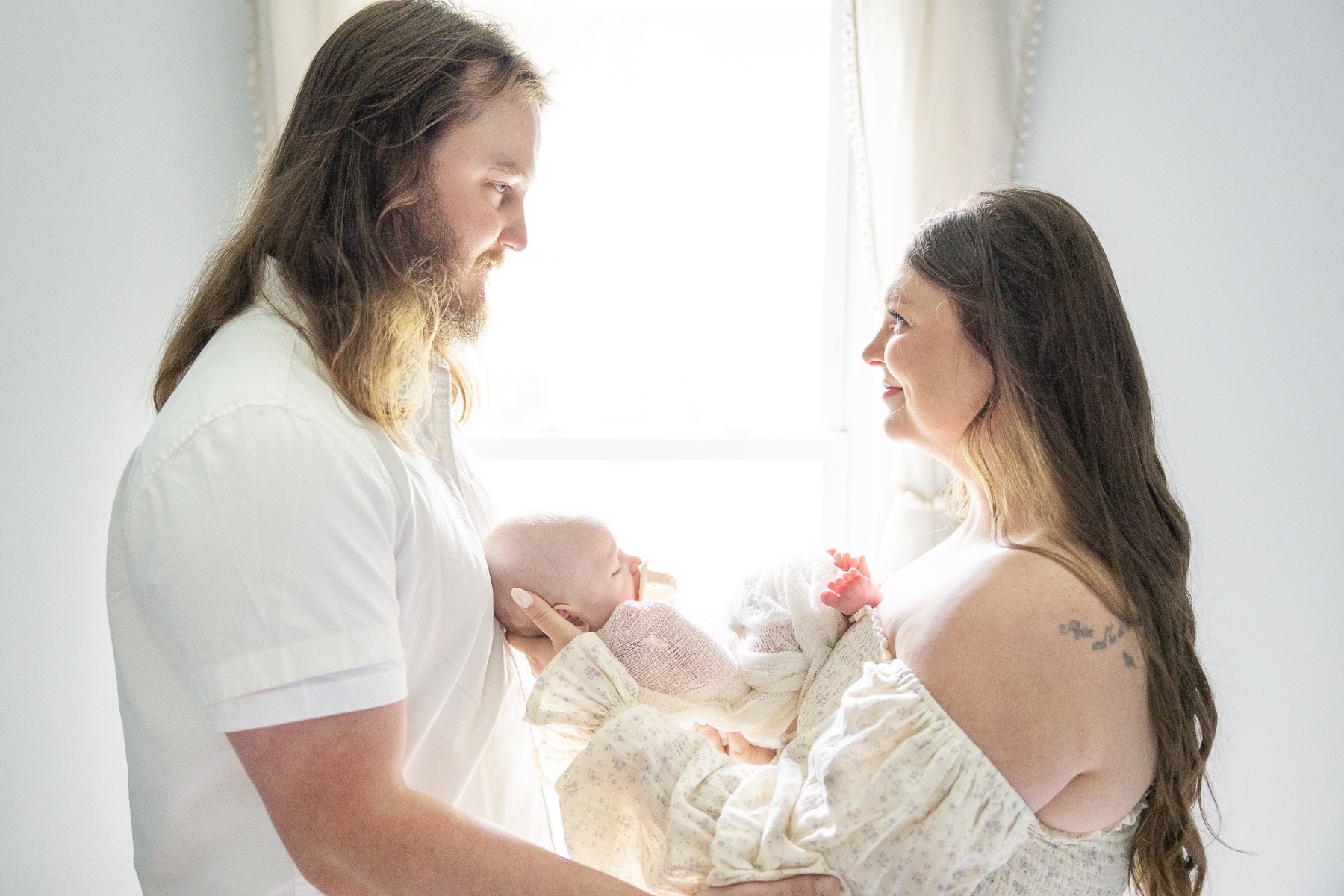 mother holds newborn between her and her husband as they look at eachother, lifestyle newborn photography
