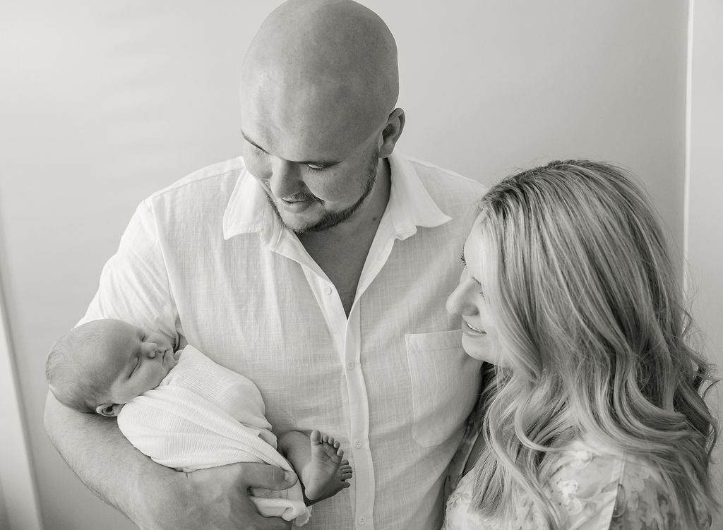 A father in a white shirt holds his newborn in his arm while mom leans into his other side