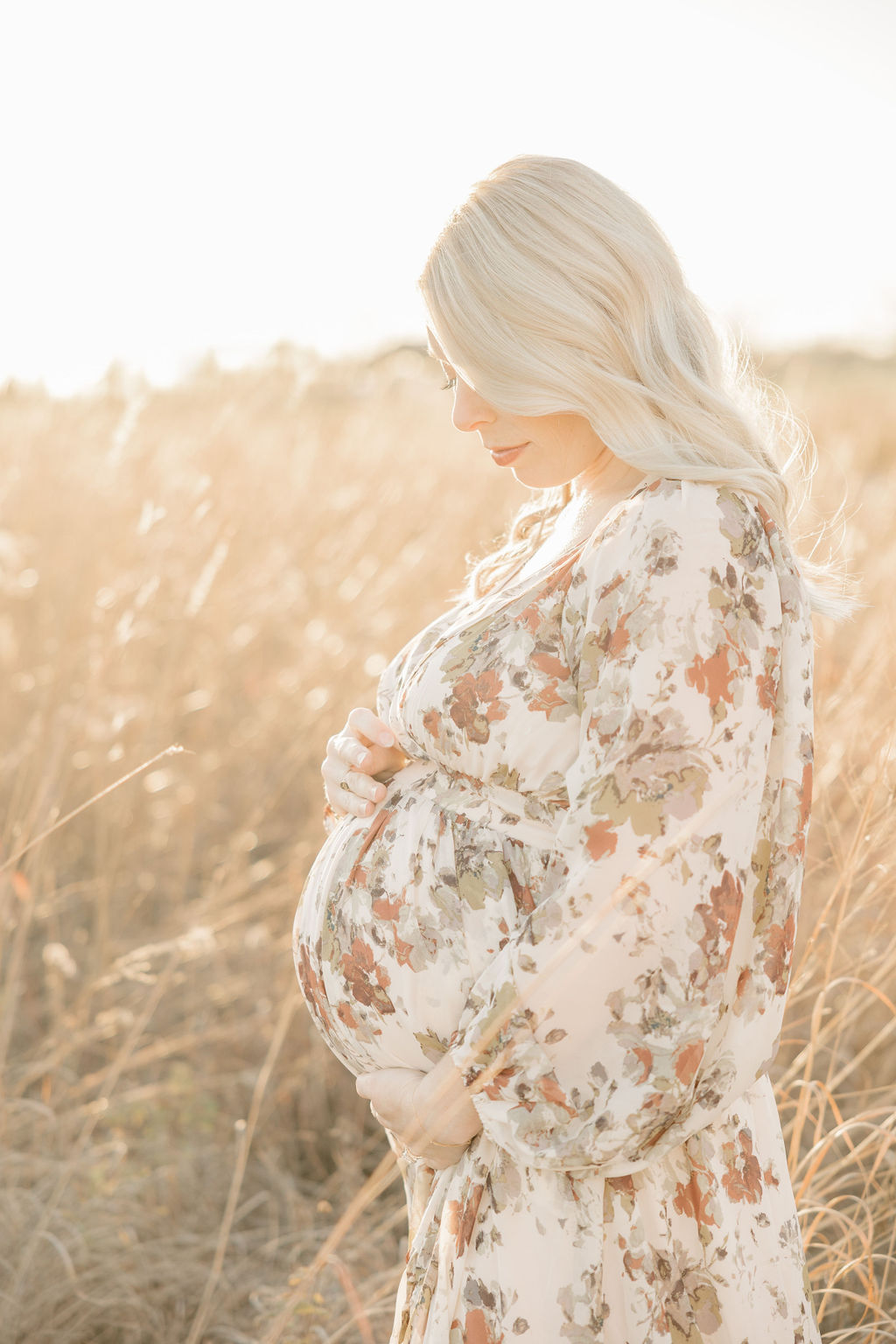 A mom to be in a white floral dress stands in a field of tall golden grass at sunset while looking down at her bump prenatal chiropractor indianapolis