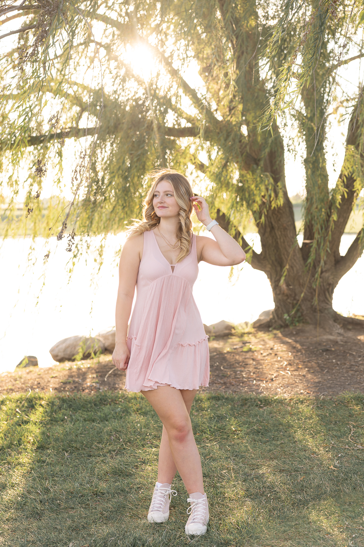 Senior girl tucks hair behind her ear in a pink dress in front of a willow tree at sunset, Carmel Senior pictures