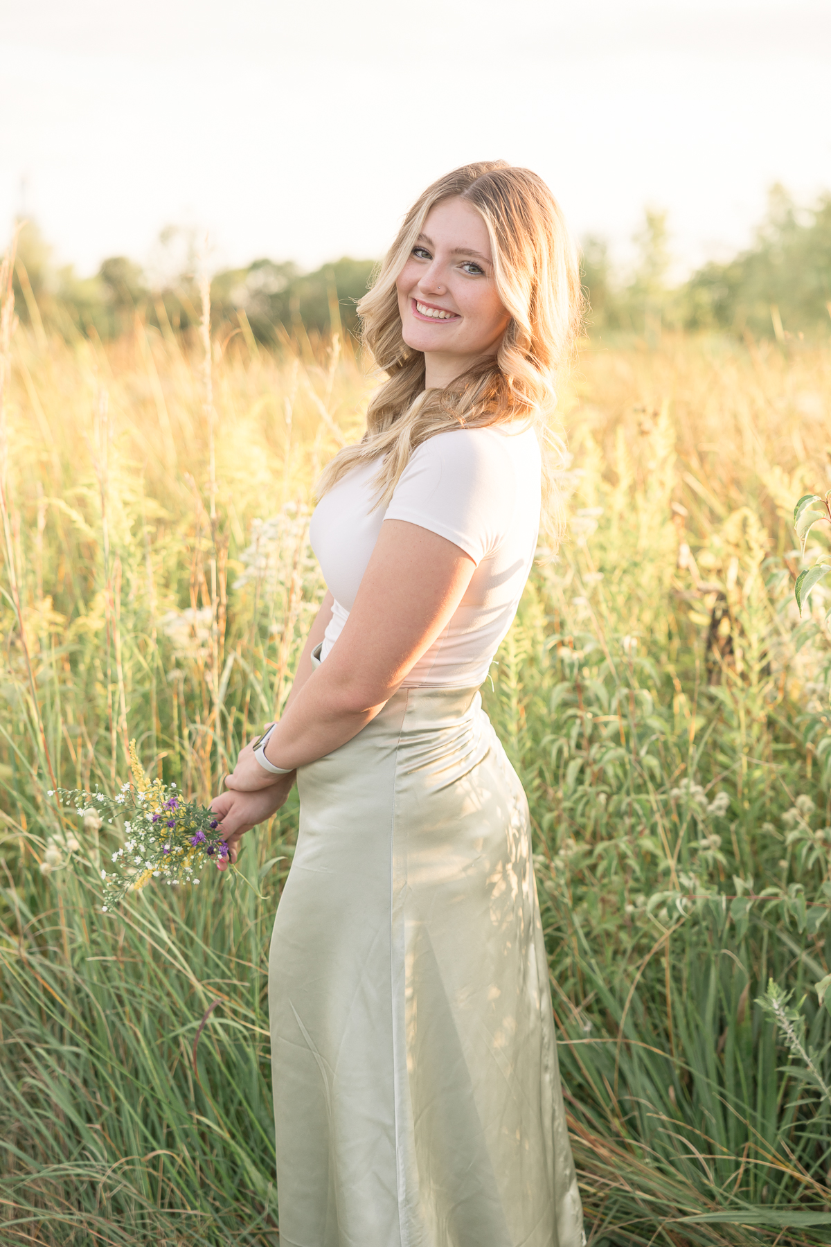 Girl in cream top and green skirt stands in field of summer grass at sunset, carmel senior pictures 