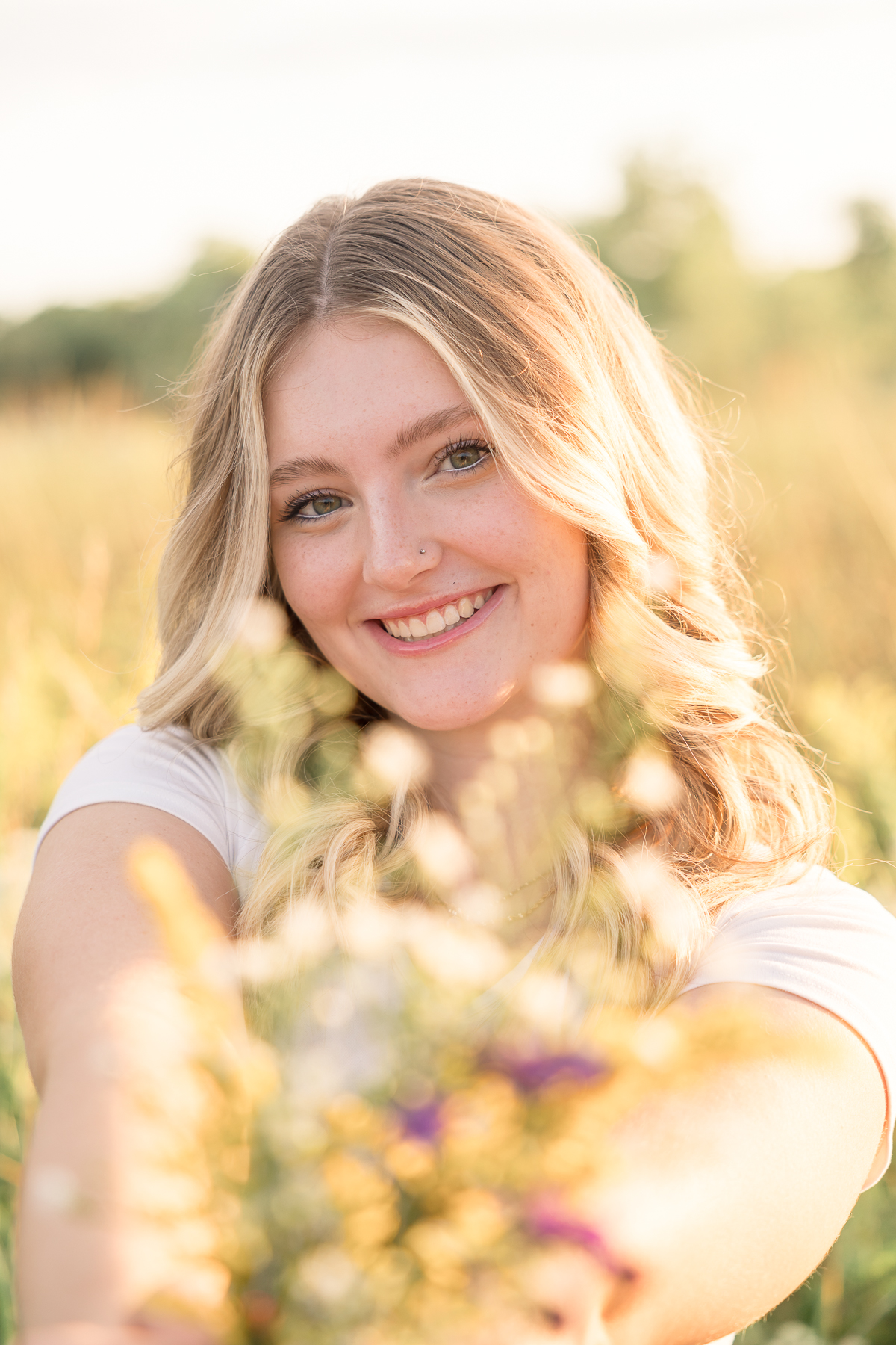 Girl holds flowers towards camera and smiles in a summer field at sunset, Carmel Senior Pictures
