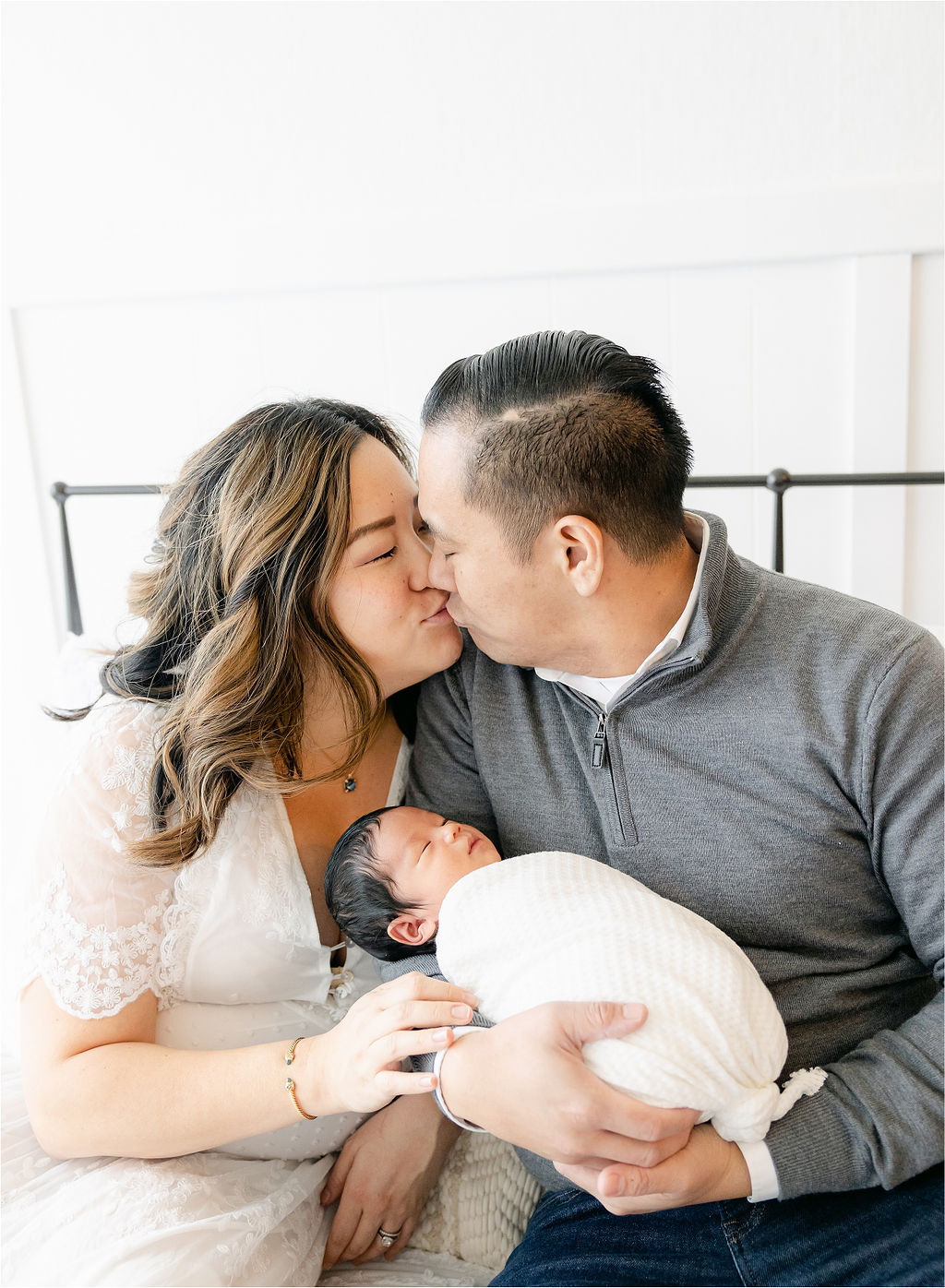 Mom and dad kiss while their newborn baby sleeps in dad's hands while sitting on a bed maternal fetal medicine indianapolis