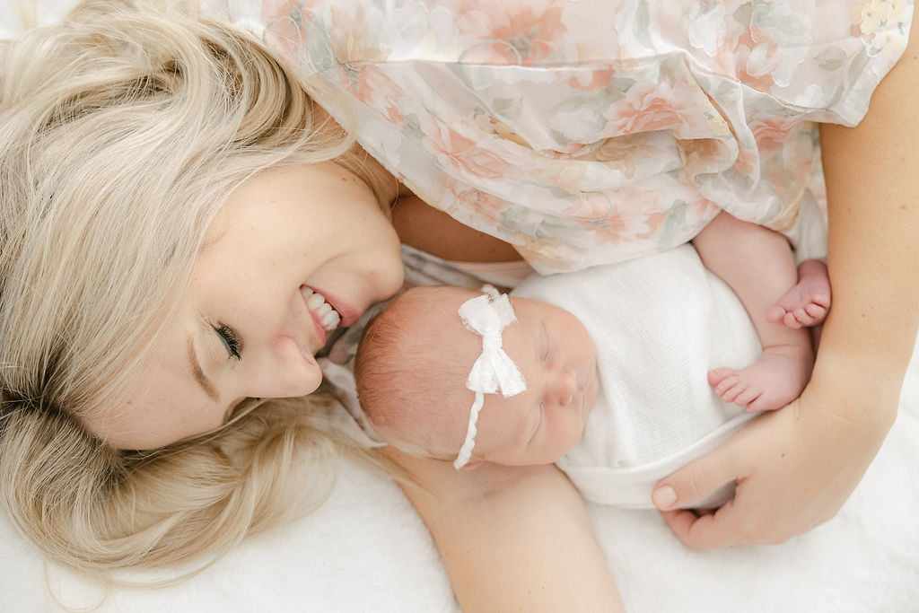 A mother in a pink floral dress cradles her sleeping newborn baby on a bed noblesville obgyn