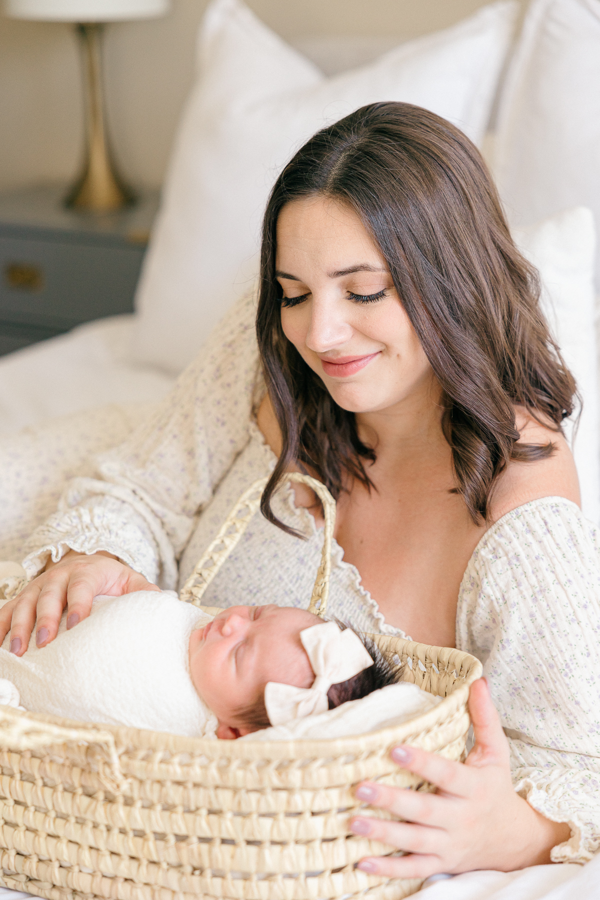 New mother looks down at her baby girl with cream bow sleeps in a moses basket, Fishers Newborn Photographer