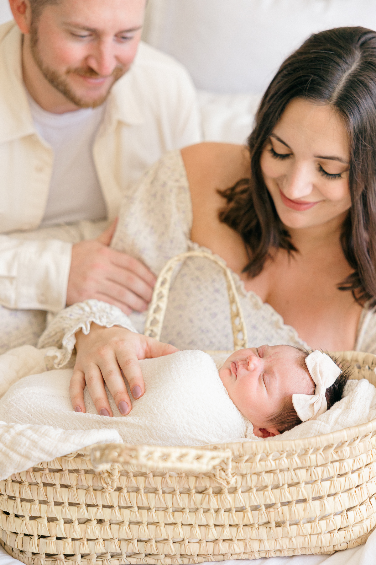 New parents look lovingly at their baby girl swaddled in cream blanket sleeps in a moses basket, Fishers Newborn Photographer
