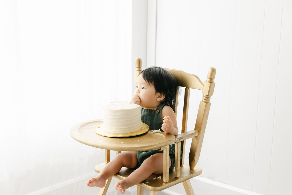 A young toddler sits in a gold high chair while wearing green overalls and eating cake